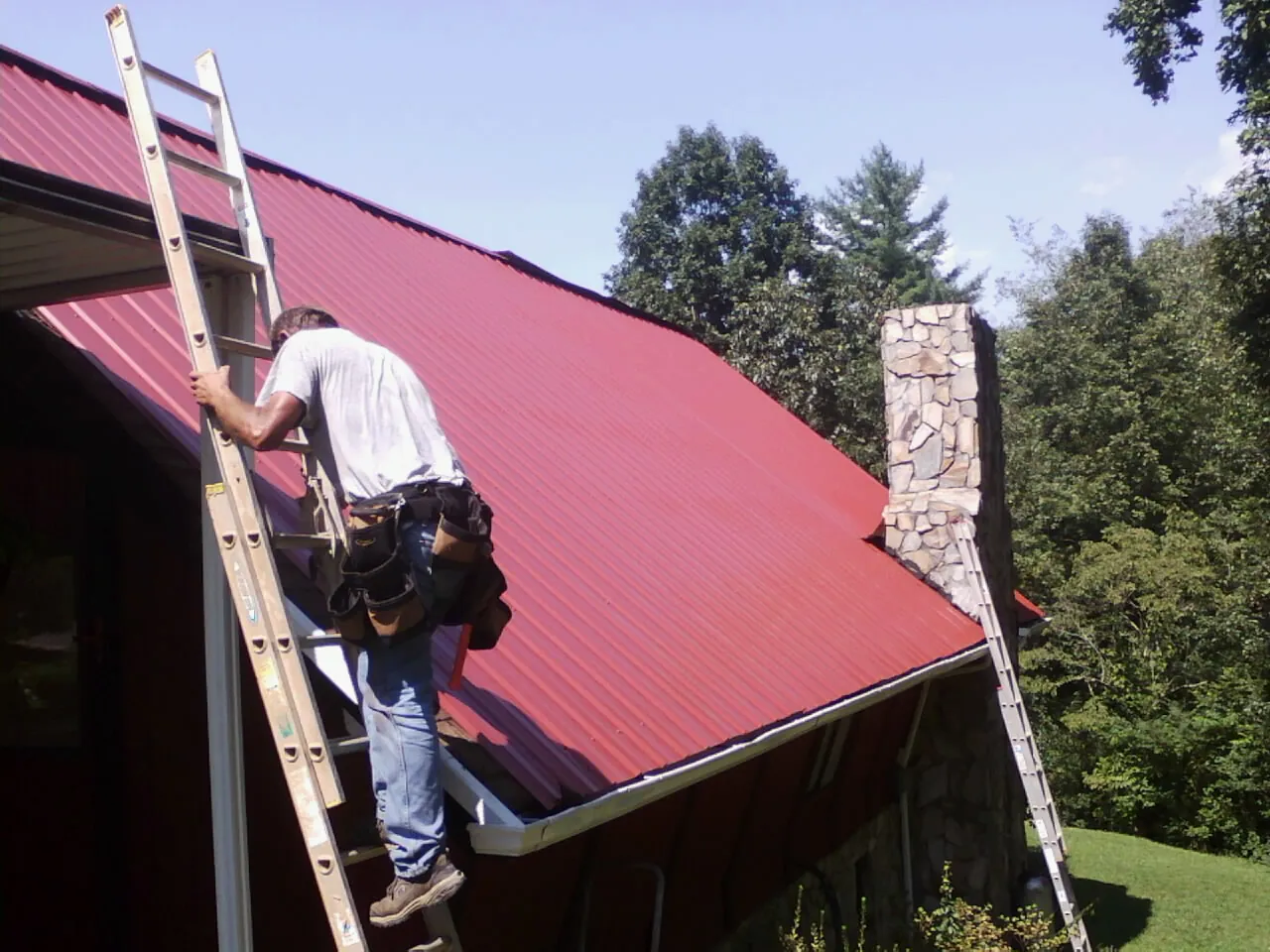 Roofer on a ladder beside a red metal roof and stone chimney — Soffit & Fascia Repair by Roofing Brothers in Silvis